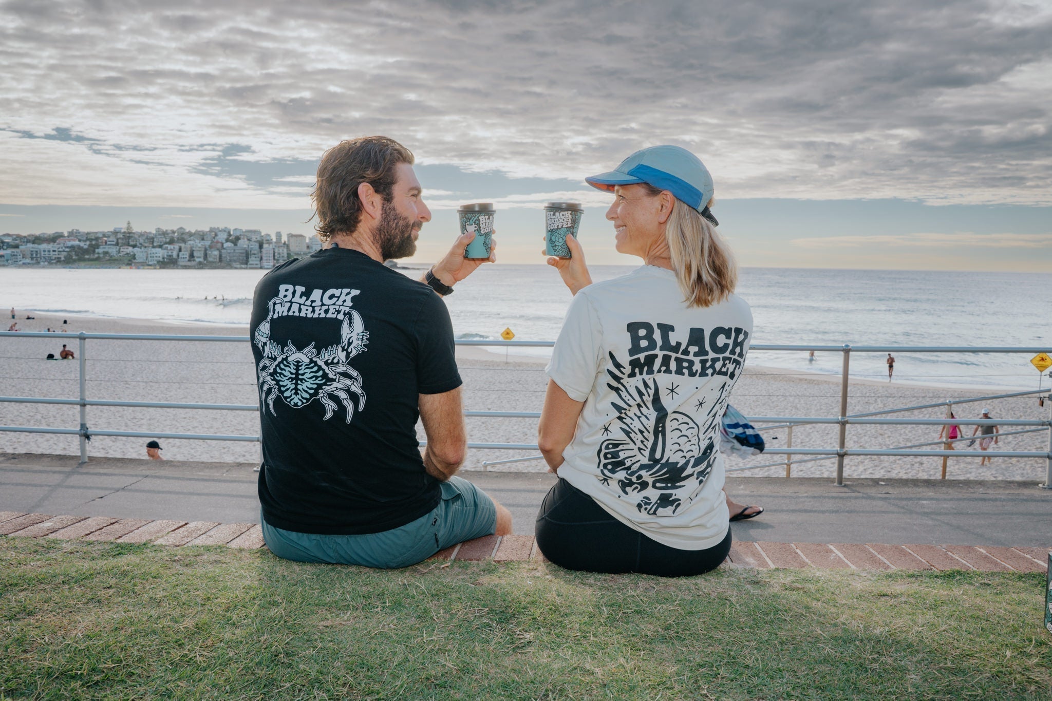 Two people sitting at Bondi beach holding a black market coffee coffee cup